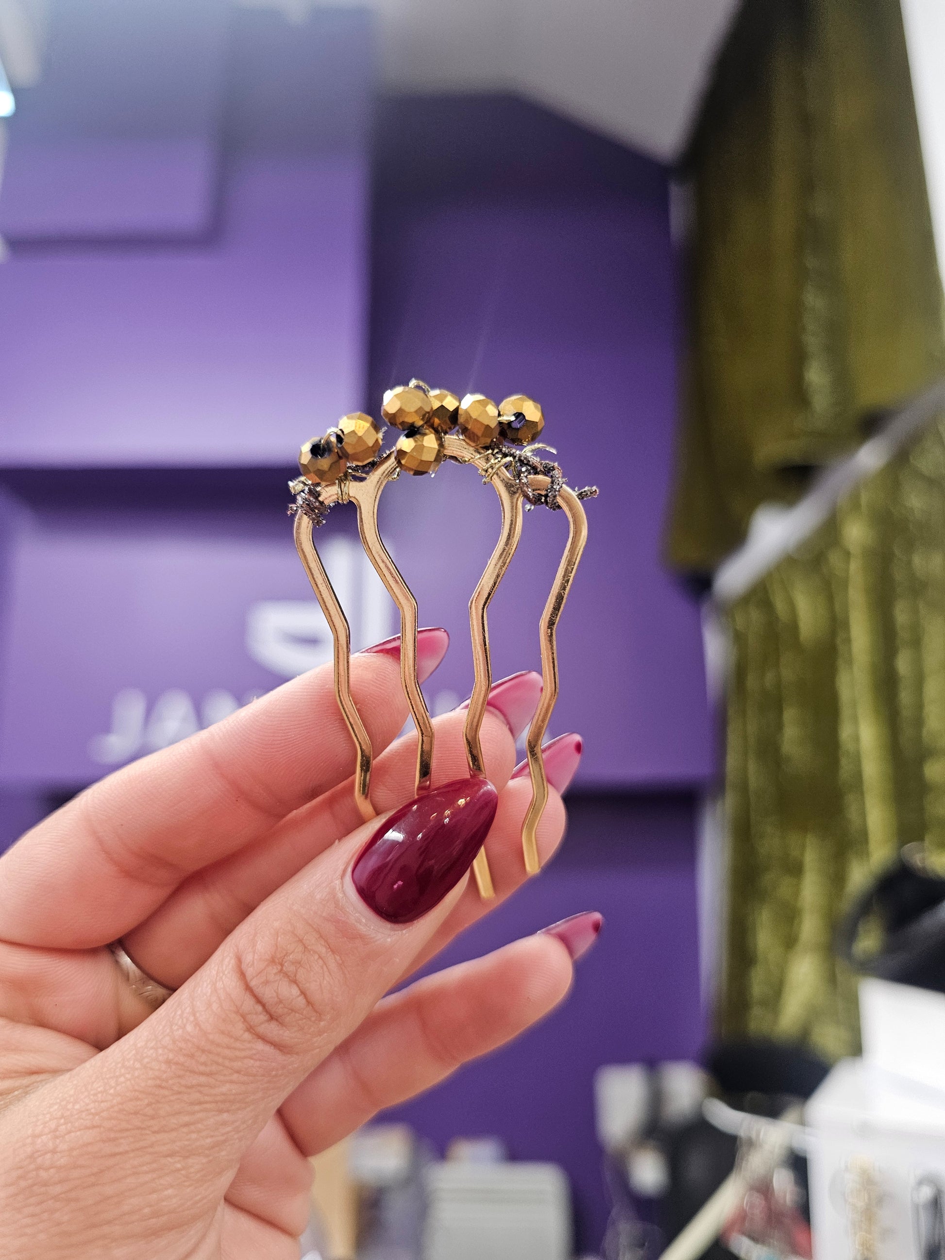 Gold and black hair clip held by a hand with red nail polish against a janizuka background