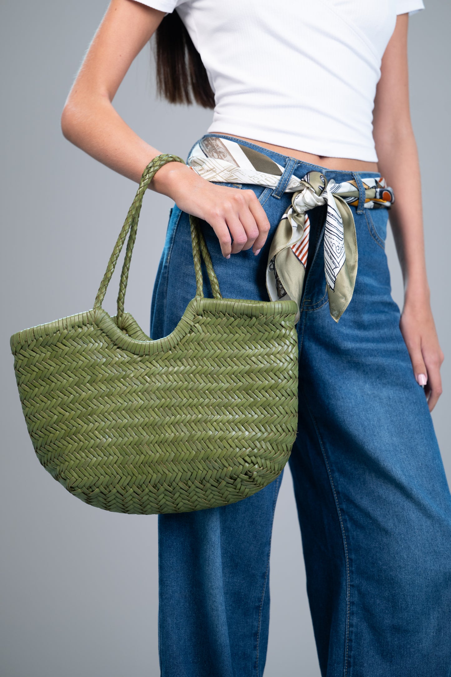 Model styling a 100% mulberry silk square scarf (olive/black/marigold print) as a belt with blue jeans, holding a green woven tote