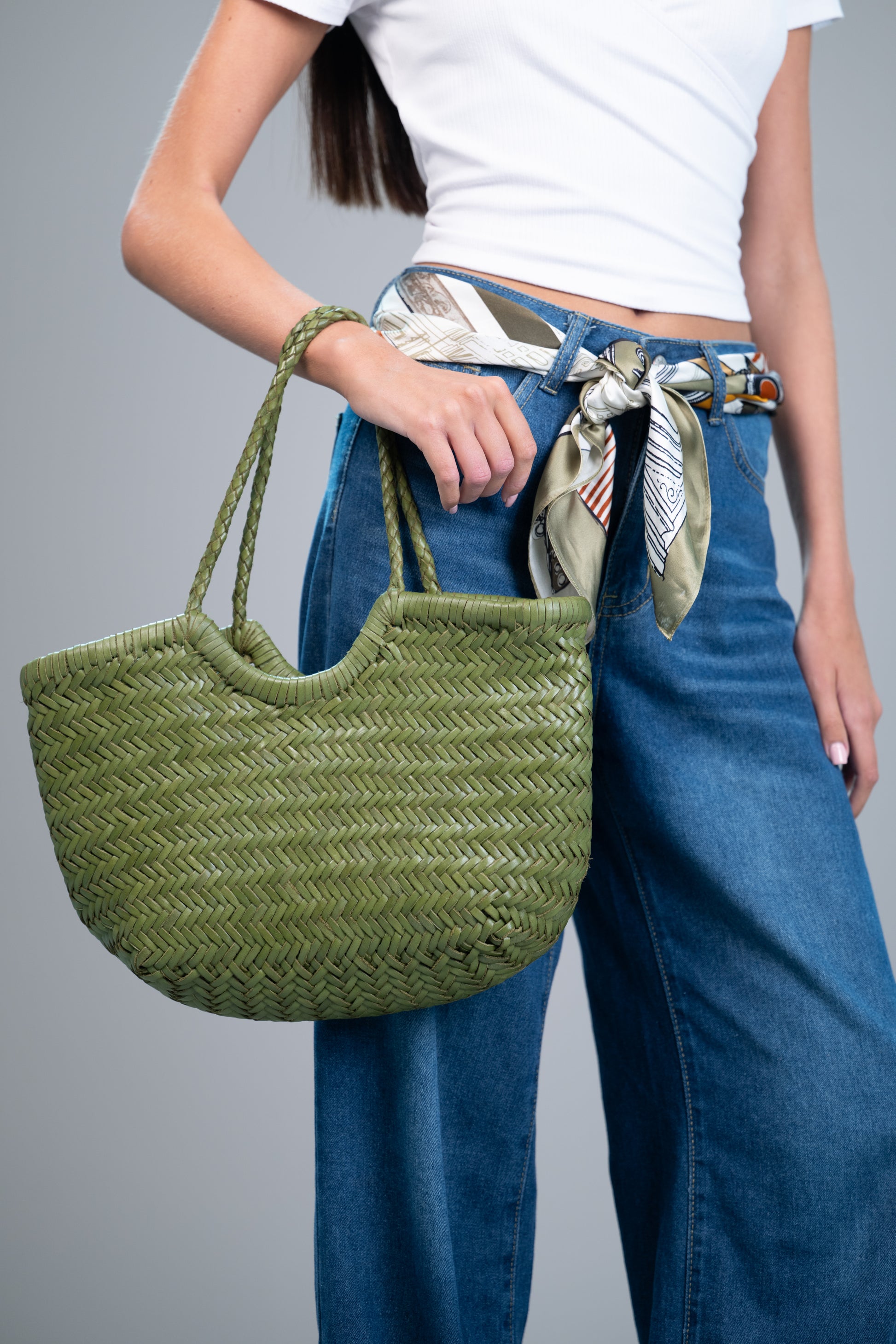 Model styling a 100% mulberry silk square scarf (olive/black/marigold print) as a belt with blue jeans, holding a green woven tote